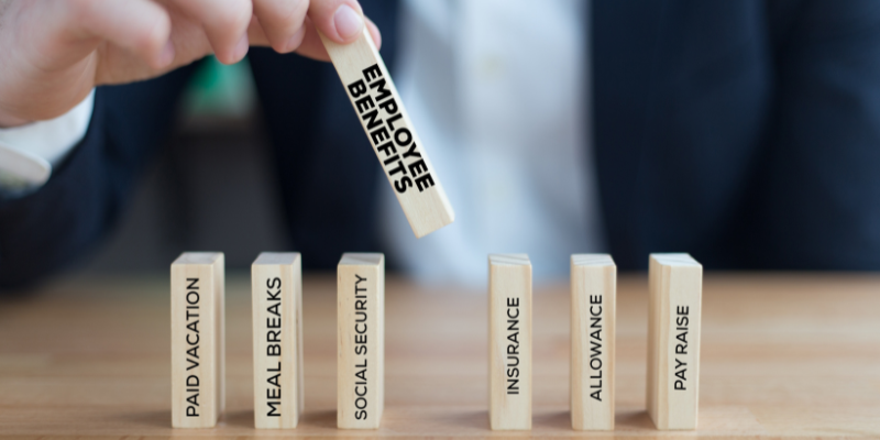 Image shows a hand holding a wooden block with the words "employee benefits", with other blocks in the foreground carved with various employee benefits