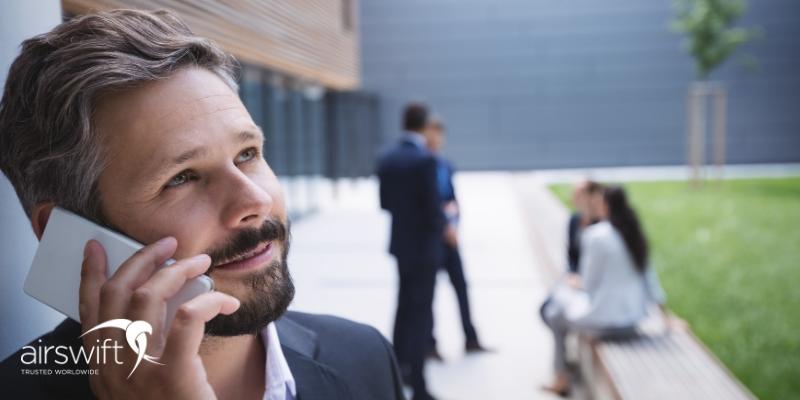 A man in professional attire on his phone, suggesting that he is leaving a professional voice message, with his colleagues in the background