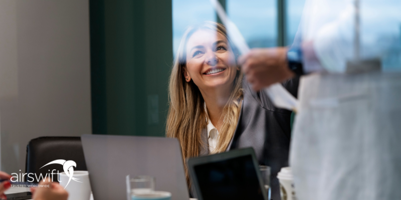 A woman in professional attire sitting at a conference table while looking up and smiling, listening to her colleague who is standing with some documents in his hands A woman in professional attire sitting at a conference table while looking up and smiling, listening to her colleague who is standing with some documents in his hands