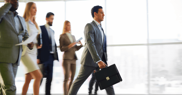 A man in a suit, holding a briefcase, walks ahead of other people who are also dressed up in business wear. 