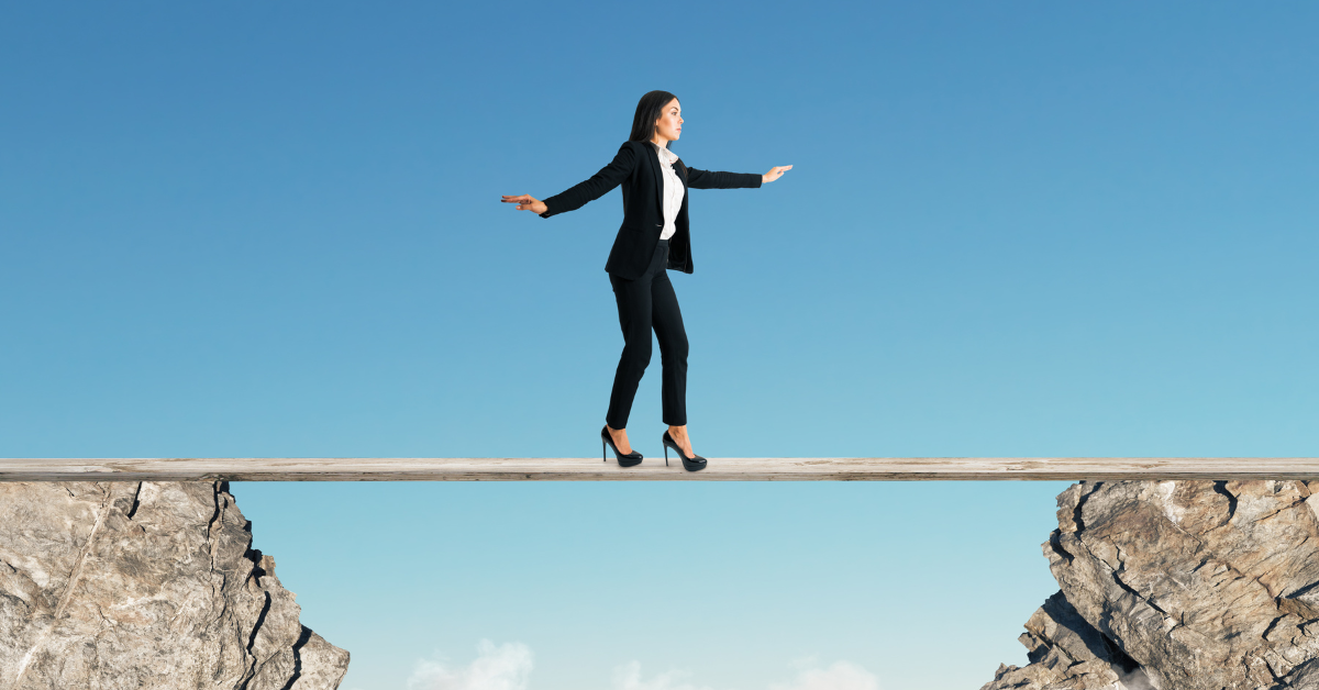 A woman in a business suit balancing on a plank between two edges of a ravine.