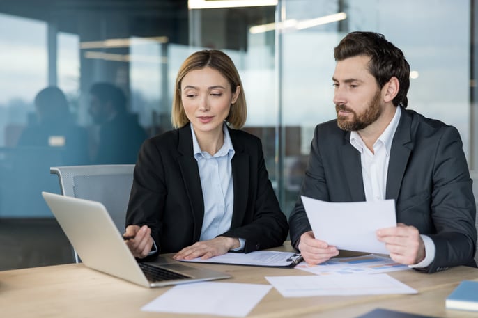 Two professionals in business attire having a meeting in an office with their laptop open and papers.