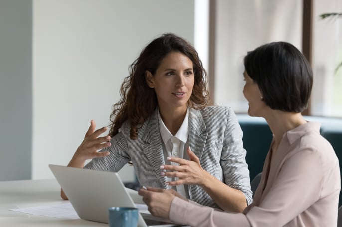 Two female professionals sat next to each other having a meeting with their laptop open and smiling mid conversation