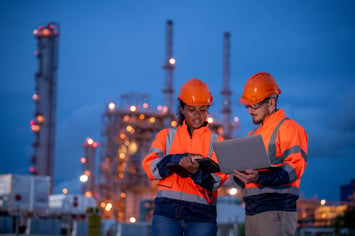 Oil and gas workers reviewing plans on laptop and clipboard at refinery site with safety gear and industrial background