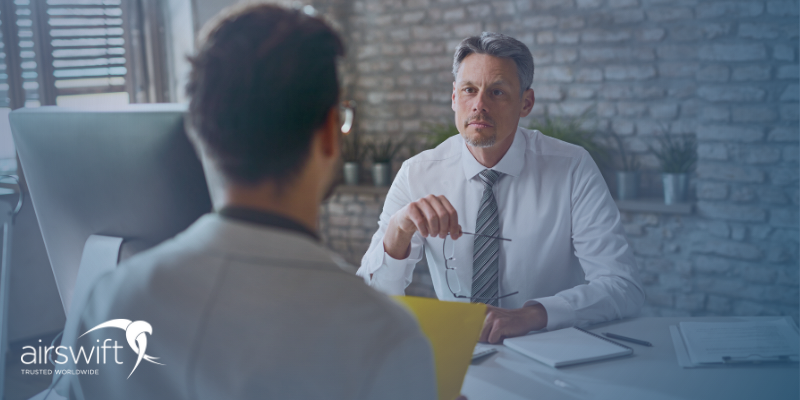 A man sits in a modern office, interviewing a male candidate for a job