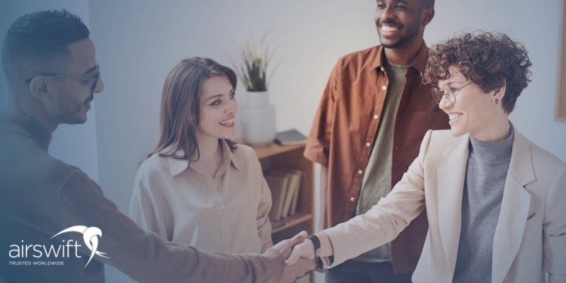 Three people (two men, one female) smile at a woman who has just completed a successful interview; one of them men shakes her hand