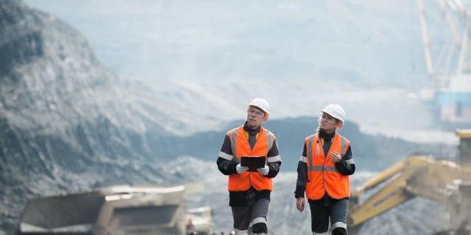 A man and woman in orange hi-vis and hardhats carrying clipboard at a mine