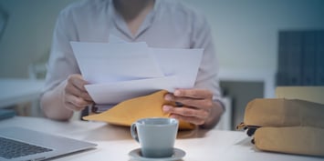 A person holding a retirement letter over an envelope, with a laptop and a cup of coffee on a desk in a softly lit office space.