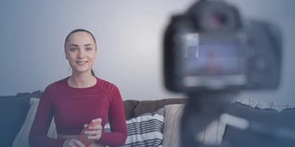 A young woman wearing a red sweater sits on her sofa in front of a camera. She is recording a video. 