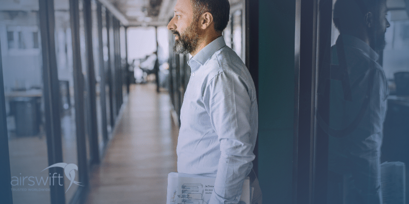 A man stands in an office corridor, looking into a glass office space