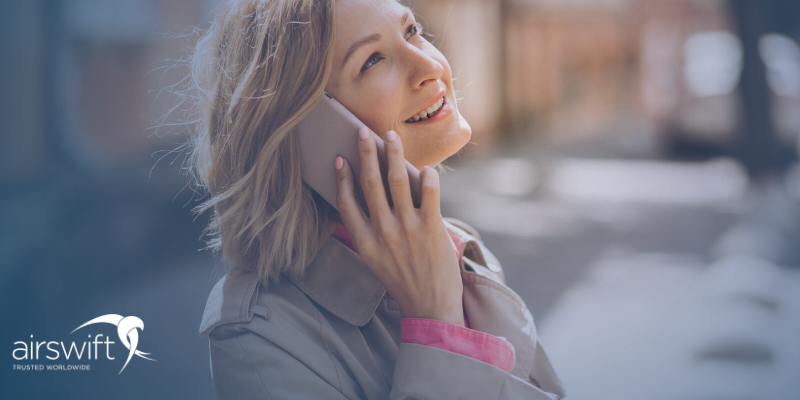 A blonde woman wearing a coat smiles as she speaks on her mobile phone. She is outside an office building