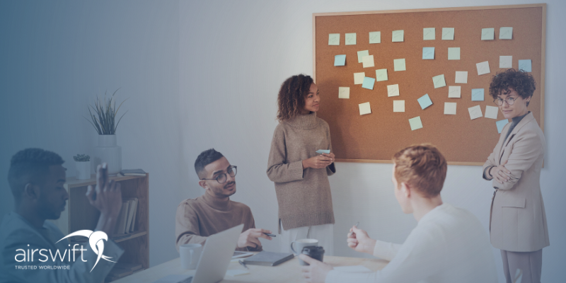 A group of colleagues sit at a table, while two women present ideas on a cork board