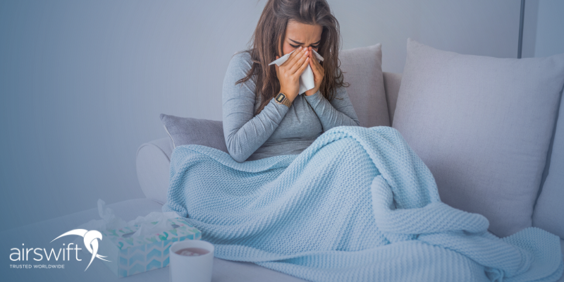 A woman sits on her sofa blowing her nose. She is wrapped in a blue blanket