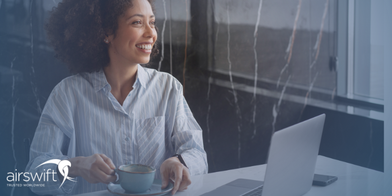 A woman smiles at her desk, with a cup of tea, her laptop open in front of her