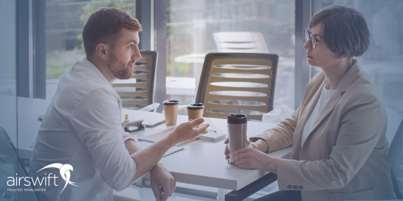 A male employee sits in a discussion with a female manager over cups of coffee