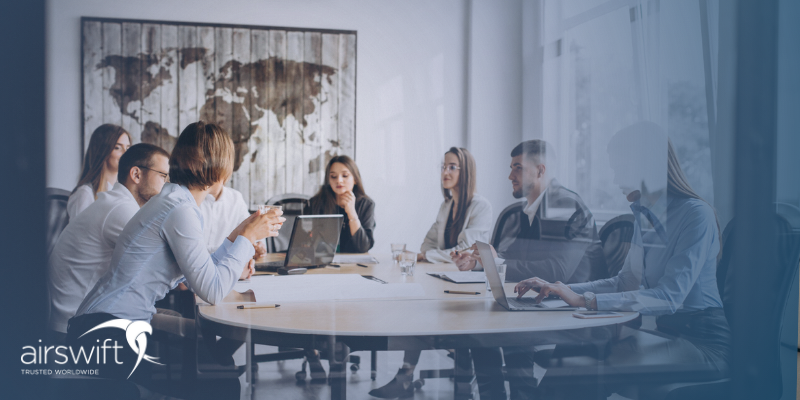 a group of professionals sit around a desk in a glass office, having a meeting around a laptop a group of professionals sit around a desk in a glass office, having a meeting around a laptop