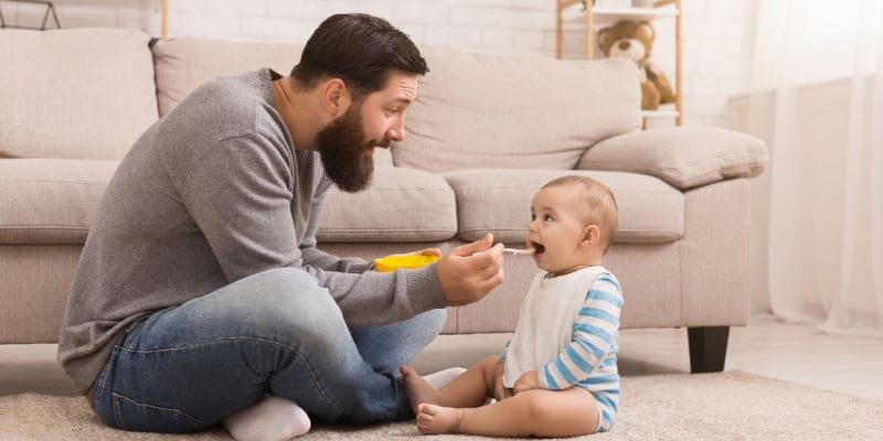 Father feeding his infant child whilst seated on the floor in their home
