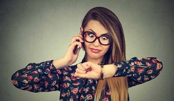 A lady in spectacles with a mobile phone to her ear and looking at her watch, indicating she is waiting for someone who is late to a meeting