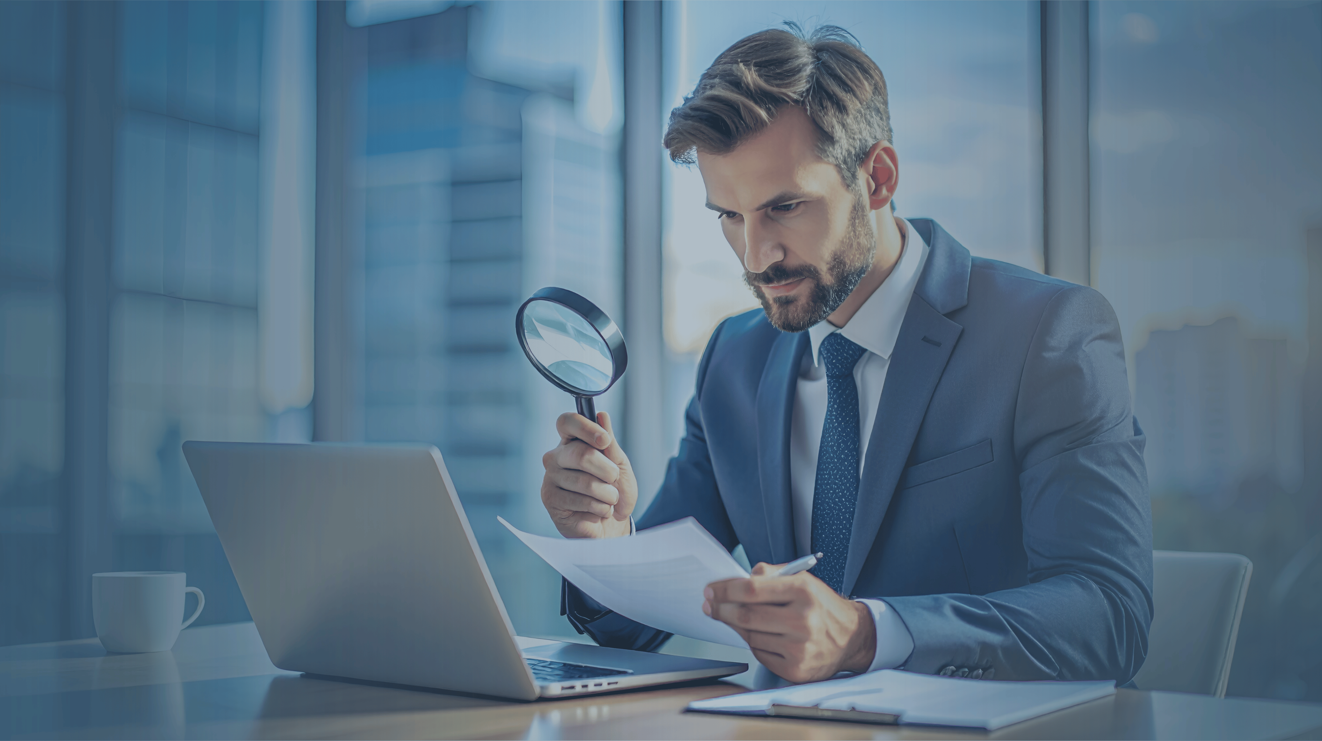 Man in office in a suit sitting at desk with laptop open using a magnifying glass and holding papers