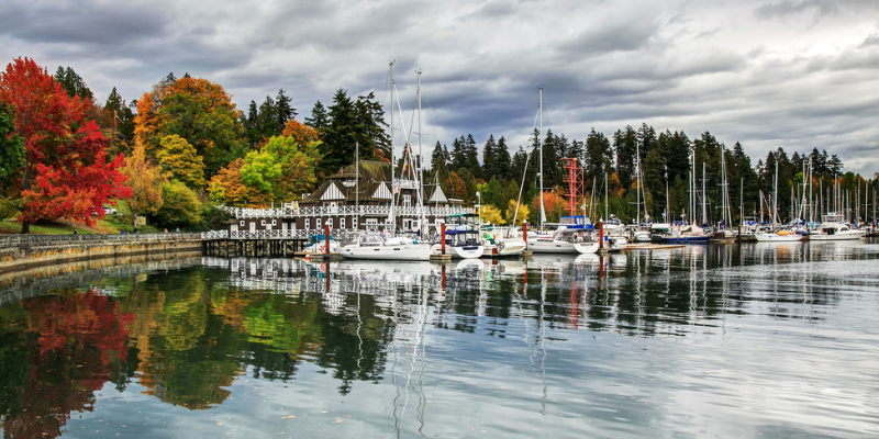 Photo shows a panoramic shot of Stanley Park in Canada featuring boats parked on a lake surrounded by autumnal trees and a cloudy overcast sky