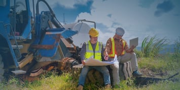 Two men wearing hard hats examine blueprints while seated on a log, discussing plans for a project.
