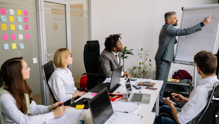 People in business attire around a conference table paying attention to a man presenting at the head of the table