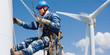 Wind turbine technician wearing safety gear and climbing equipment works on a turbine tower with another turbine visible in the background under a clear blue sky. This image illustrates one of the fastest growing jobs in the U.S. and highlights high demand positions in the country..