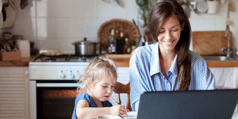 Woman working at her desk with her laptop open in fron of her and her toddler daugther is sitting to her left. Background is of her kitchen.