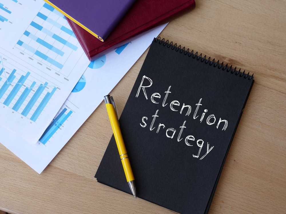 (Foreground) A spiral notebook with the words "retention strategy" on the cover, with a yellow pen on it. (Background) Other notebooks and some printouts of charts.