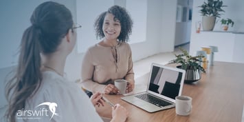 Two professionals engaged in an informal interview conversation in a modern office, with one person smiling and holding a coffee mug while the other takes notes.
