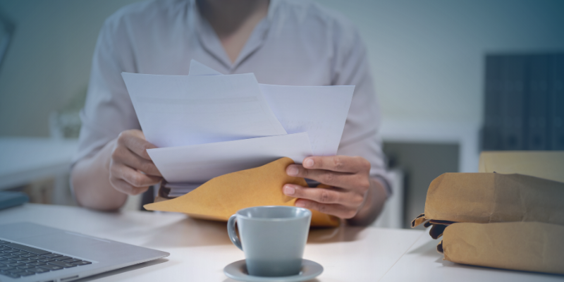 A person holding a retirement letter over an envelope, with a laptop and a cup of coffee on a desk in a softly lit office space.