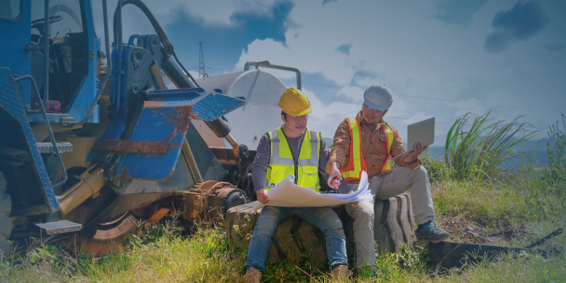 Two men wearing hard hats examine blueprints while seated on a log, discussing plans for a project.